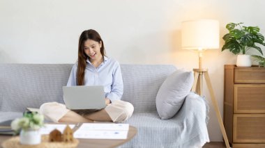 Woman using laptop to work or do homework at home with smiling face in her living room, Creating happiness at work with a smile, Live performance or video call with laptop, Work from home.