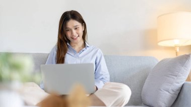 Woman using laptop to work or do homework at home with smiling face in her living room, Creating happiness at work with a smile, Live performance or video call with laptop, Work from home.