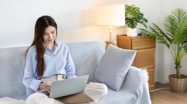 Woman using laptop to work or do homework at home with smiling face in her living room, Creating happiness at work with a smile, Live performance or video call with laptop, Work from home.