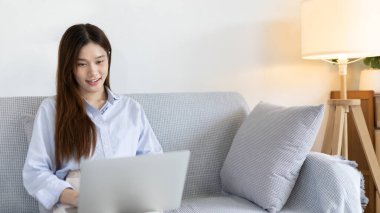 Woman using laptop to work or do homework at home with smiling face in her living room, Creating happiness at work with a smile, Live performance or video call with laptop, Work from home.