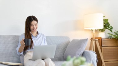 Beautiful Asian woman working on laptop and sipping coffee with smiling face in her home, Creating happiness at work with a smile, Freelancer working at home happily, Work from home.