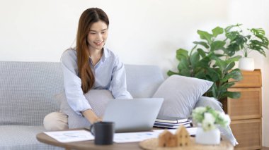 Woman using laptop to work or do homework at home with smiling face in her living room, Creating happiness at work with a smile, Live performance or video call with laptop, Work from home.
