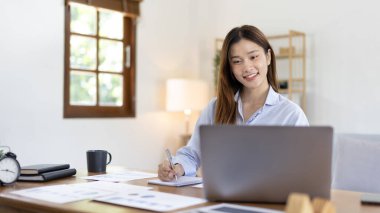 Beautiful young asian woman watching live video or video call of teacher teaching on laptop in her home, Take notes of important conversations and messages during the teacher's teaching.