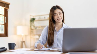 Beautiful young asian woman watching live video or video call of teacher teaching on laptop in her home, Take notes of important conversations and messages during the teacher's teaching.