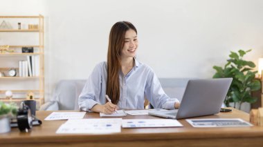 Beautiful young asian woman watching live video or video call of teacher teaching on laptop in her home, Take notes of important conversations and messages during the teacher's teaching.