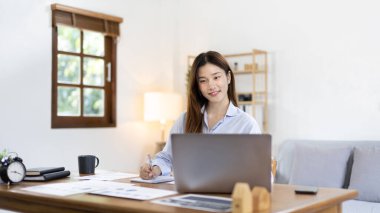 Beautiful young asian woman watching live video or video call of teacher teaching on laptop in her home, Take notes of important conversations and messages during the teacher's teaching.