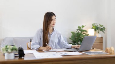 Beautiful young asian woman watching live video or video call of teacher teaching on laptop in her home, Take notes of important conversations and messages during the teacher's teaching.