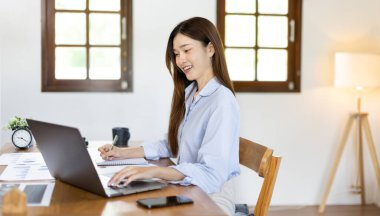 Beautiful young asian woman watching live video or video call of teacher teaching on laptop in her home, Take notes of important conversations and messages during the teacher's teaching.