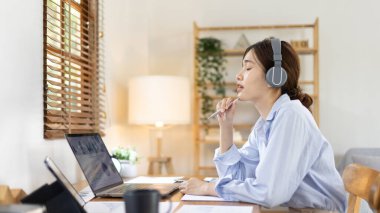 Asian female college student wearing headphones watching live performance or video call teacher teaching on laptop, Conversations with teachers and classmates, Online learning, Study at your own home.