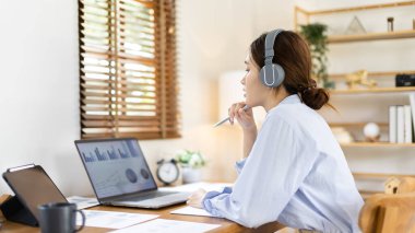 Asian female college student wearing headphones watching live performance or video call teacher teaching on laptop, Conversations with teachers and classmates, Online learning, Study at your own home.