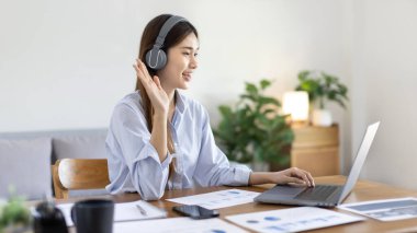 Asian female college student wearing headphones watching live performance or video call teacher teaching on laptop, Conversations with teachers and classmates, Online learning, Study at your own home.