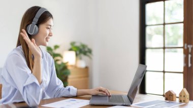 Asian female college student wearing headphones watching live performance or video call teacher teaching on laptop, Conversations with teachers and classmates, Online learning, Study at your own home.