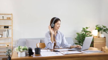 Asian female college student wearing headphones watching live performance or video call teacher teaching on laptop, Conversations with teachers and classmates, Online learning, Study at your own home.