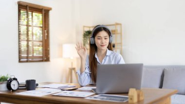 Asian female college student wearing headphones watching live performance or video call teacher teaching on laptop, Conversations with teachers and classmates, Online learning, Study at your own home.