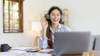 Asian female college student wearing headphones watching live performance or video call teacher teaching on laptop, Conversations with teachers and classmates, Online learning, Study at your own home.
