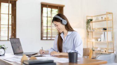 Asian female college student wearing headphones watching live performance or video call teacher teaching on laptop, Conversations with teachers and classmates, Online learning, Study at your own home.
