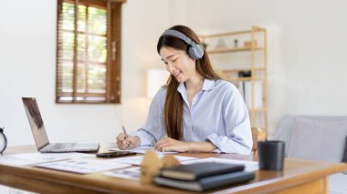 Asian female college student wearing headphones watching live performance or video call teacher teaching on laptop, Conversations with teachers and classmates, Online learning, Study at your own home.