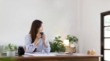 Beautiful Asian woman working on laptop and sipping coffee with smiling face in her home, Creating happiness at work with a smile, Freelancer working at home happily, Work from home.