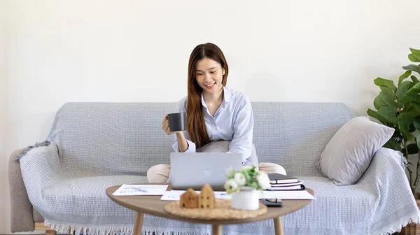 Beautiful Asian woman working on laptop and sipping coffee with smiling face in her home, Creating happiness at work with a smile, Freelancer working at home happily, Work from home.
