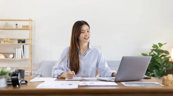 Beautiful young asian woman watching live video or video call of teacher teaching on laptop in her home, Take notes of important conversations and messages during the teacher\'s teaching.