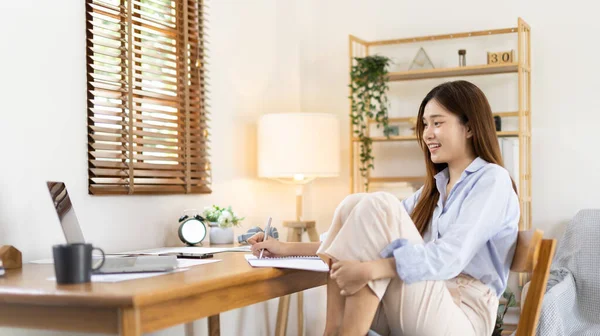Beautiful young asian woman watching live video or video call of teacher teaching on laptop in her home, Take notes of important conversations and messages during the teacher's teaching.