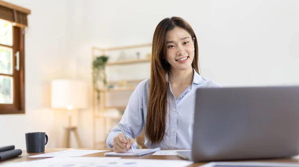 Beautiful young asian woman watching live video or video call of teacher teaching on laptop in her home, Take notes of important conversations and messages during the teacher\'s teaching.