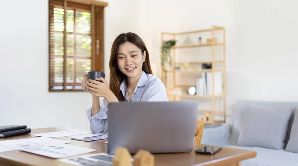 Beautiful Asian woman working on laptop and sipping coffee with smiling face in her home, Creating happiness at work with a smile, Freelancer working at home happily, Work from home.