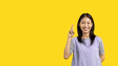 Young Asian woman making a symbolic gesture with fingers crossed showing good luck, White lie gesture, Fingers crossed, Woman doing hand sign on yellow background, Superstitious concept.