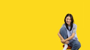 Asian biracial woman sitting in wooden chair and smiling happily on yellow background, Lovely lonely woman spreads a bright smile, Smile is cheerful and bright.