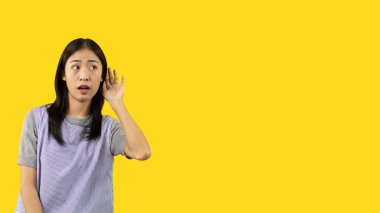Asian woman eavesdropping or overhearing secret conversation isolated on yellow background, Gossip, Listening, Yellow background studio portrait.