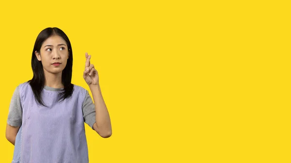 Young Asian woman making a symbolic gesture with fingers crossed showing good luck, White lie gesture, Fingers crossed, Woman doing hand sign on yellow background, Superstitious concept.