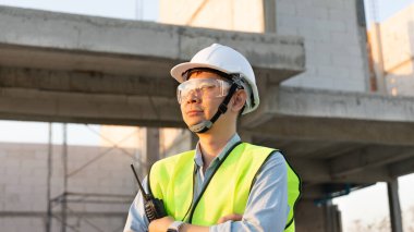 Usta mühendis fabrika inşaatını inceliyor ve sahada çalışan meslektaşlarıyla sohbet etmek için bir telsiz kullanıyor, mühendis dışarıda planlarla çalışıyor ve koruyucu güvenlik donanımı takıyor..