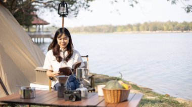 Young woman camping for the weekend in the woods near the river, Enjoying Camping Holiday In Countryside, woman sitting and relaxing happily reading definitions, Young woman outdoor leisure activities.