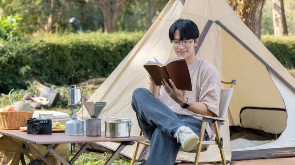 Young man camping for the weekend in the woods near the river, Camping Holiday In Countryside, Man sitting and relaxing happily reading definitions, Young man outdoor leisure activities.