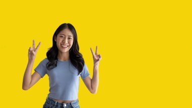 Young Asian woman making two thumbs up gesture showing joy and fun, Symbol of good friendship, Popular photo poses, Make a v-shaped finger, Yellow background.