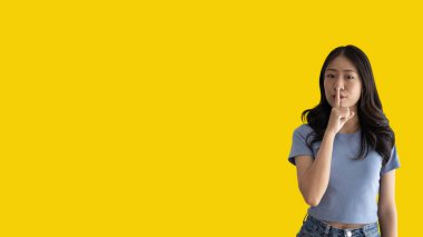 Asian woman doing silent gesture with finger, Noiseless symbol, Noisy mall, Hide a secret, Do not speak. shut your mouth, Noiseless signal transmission, Isolated on yellow background.