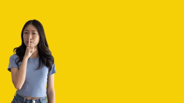 Asian woman doing silent gesture with finger, Noiseless symbol, Noisy mall, Hide a secret, Do not speak. shut your mouth, Noiseless signal transmission, Isolated on yellow background.