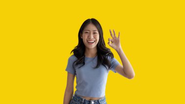 Young asian woman over yellow background doing ok sign with hands, OK symbol, Welcome, Agree, Join, Do as instructed, Follow orders, Acceptance of all conditions.