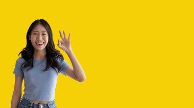 Young asian woman over yellow background doing ok sign with hands, OK symbol, Welcome, Agree, Join, Do as instructed, Follow orders, Acceptance of all conditions.