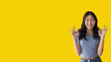 Young asian woman over yellow background doing ok sign with hands, OK symbol, Welcome, Agree, Join, Do as instructed, Follow orders, Acceptance of all conditions.