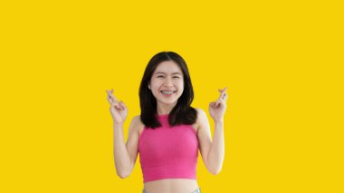 Young Asian woman making a symbolic gesture with fingers crossed showing good luck, White lie gesture, Fingers crossed, Woman doing hand sign on yellow background, Superstitious concept.