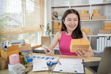 Kadın, ürün sipariş etmek için gelen müşterilerle sohbet etmek için cep telefonu kullanıyor, evde serbest iş, arka planda ambalaj, çevrimiçi satış, küçük işletme sahibi, çevrimiçi alışveriş KOBİ girişimcisi.