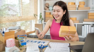 Kadın, ürün sipariş etmek için gelen müşterilerle sohbet etmek için cep telefonu kullanıyor, evde serbest iş, arka planda ambalaj, çevrimiçi satış, küçük işletme sahibi, çevrimiçi alışveriş KOBİ girişimcisi.