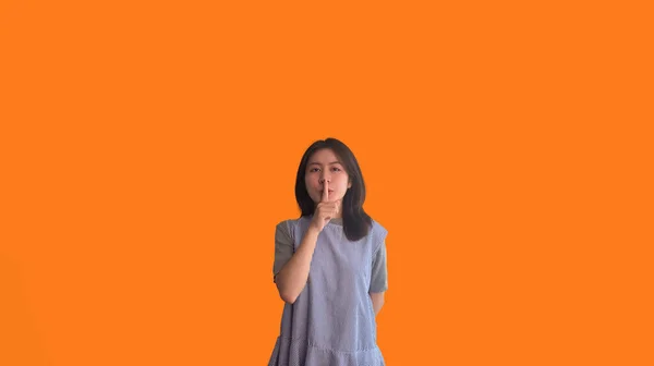 stock image Asian woman doing silent gesture with finger, Noiseless symbol, Noisy mall, Hide a secret, Do not speak. shut your mouth, Noiseless signal transmission, Isolated on orange background.