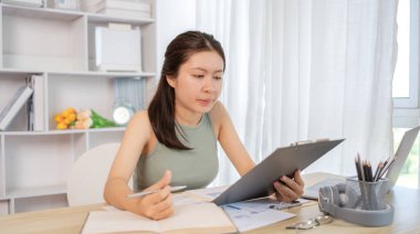 Beautiful young asian woman watching live video or video call of teacher teaching on laptop in her home, Take notes of important conversations and messages during the teacher's teaching.