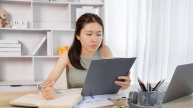 Beautiful young asian woman watching live video or video call of teacher teaching on laptop in her home, Take notes of important conversations and messages during the teacher's teaching.