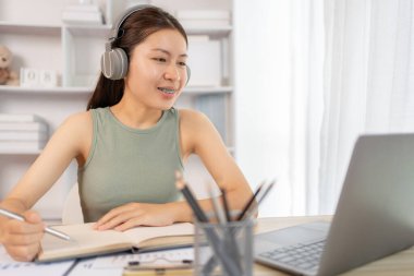 Beautiful young asian woman watching live video or video call of teacher teaching on laptop in her home, Take notes of important conversations and messages during the teacher's teaching.