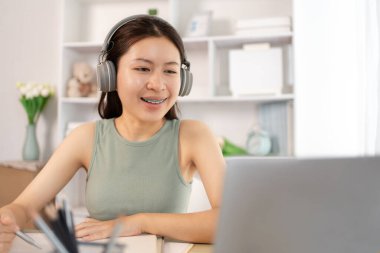 Beautiful young asian woman watching live video or video call of teacher teaching on laptop in her home, Take notes of important conversations and messages during the teacher's teaching.