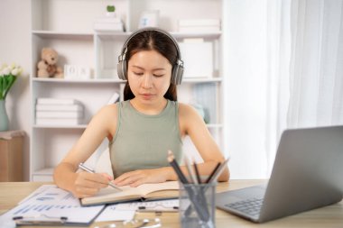 Beautiful young asian woman watching live video or video call of teacher teaching on laptop in her home, Take notes of important conversations and messages during the teacher's teaching.