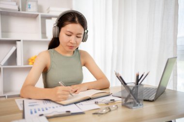 Beautiful young asian woman watching live video or video call of teacher teaching on laptop in her home, Take notes of important conversations and messages during the teacher's teaching.